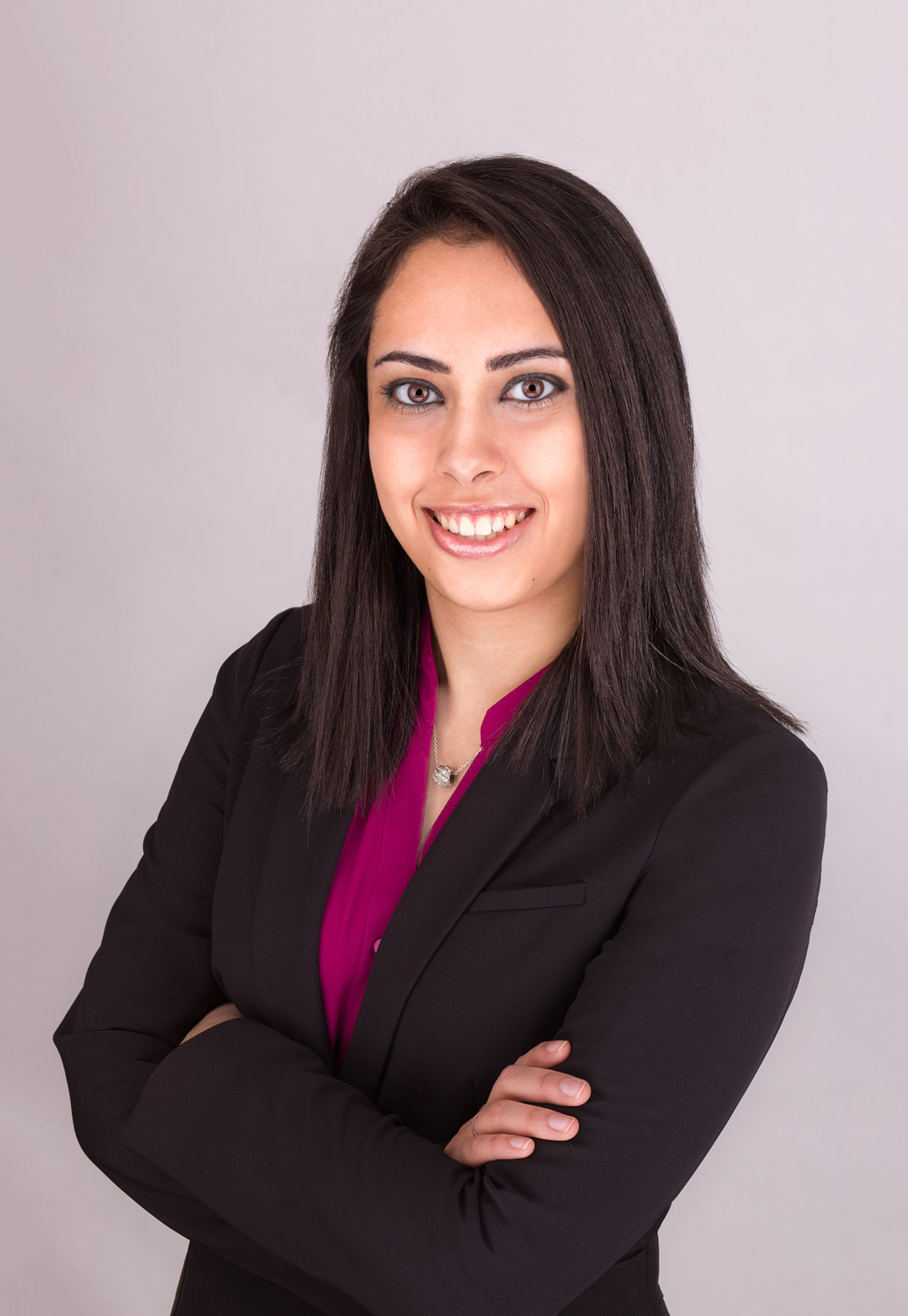 Shafagh stands with her arms crossed against a neutral background. She is smiling. She wears a smart black blazer with a dark pink shirt underneath. She has long straight dark hair and olive skin.