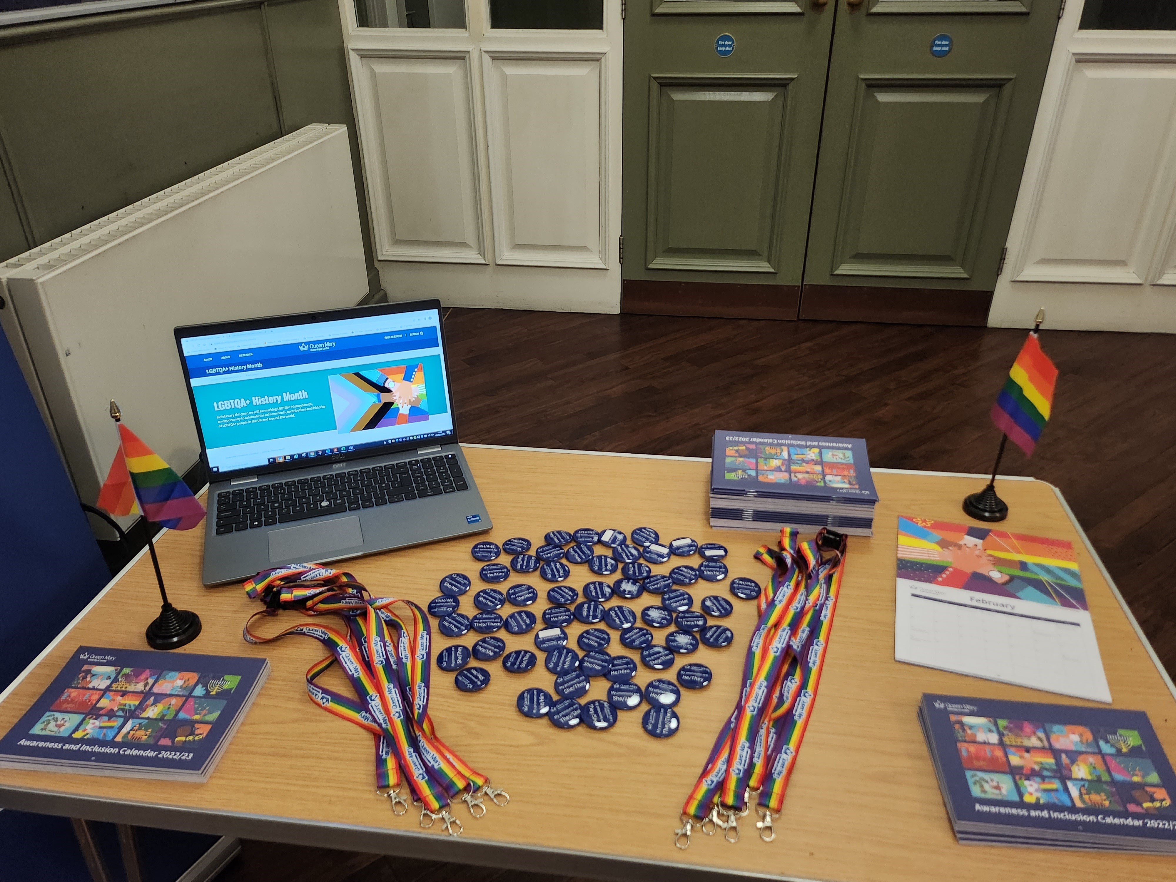Photo of a desk with Rainbow lanyards, pronoun badges, Inclusion & Awareness calendars, Pride flags and a laptop displaying the LGBTQA+ History Month webpages