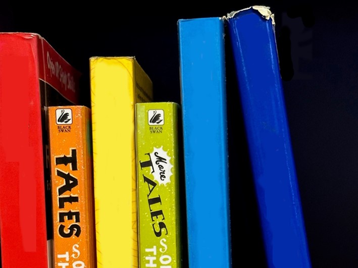 A photograph of books with multi-coloured spines, stacked horizontally, on a black background