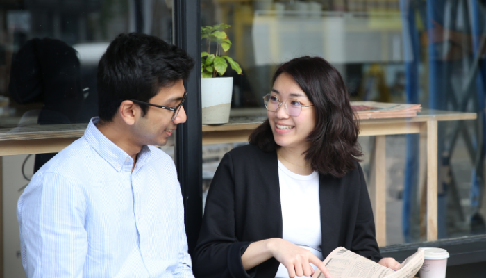 Two students sitting and talking outside a cafe one is holding a newspaper and pointing to it while smiling