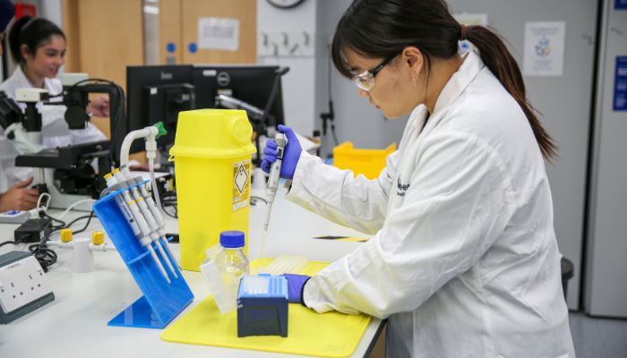 A student wearing a lab coat and protective glasses with hair in a long ponytail using equipment in a lab