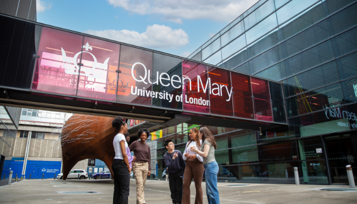A group of students dressed casually standing and talking outside and infront of buildings with Queen Mary University of London wrote on it