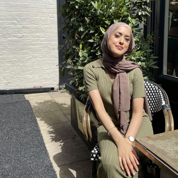 Photo of Ayeesha Miah. She is sitting in a sunny courtyard wearing a khaki dress and a brown hijab. Her hands are placed over her legs which are crossed at the knee.