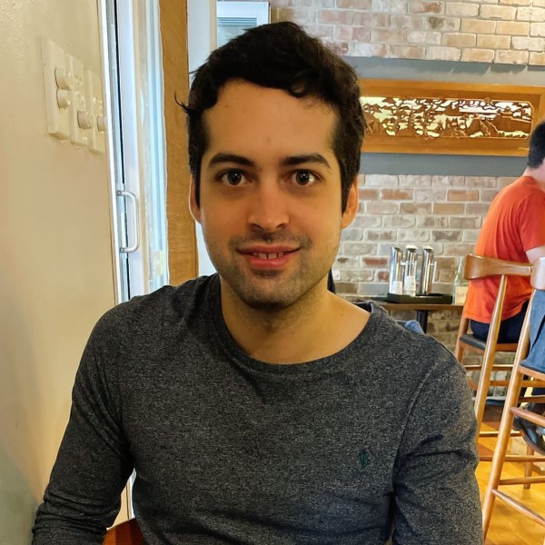 Headshot of alumnus, Luiz Pereira. He is sat in a restaurant, wearing a long-sleeved grey sweater and is looking directly at the camera.