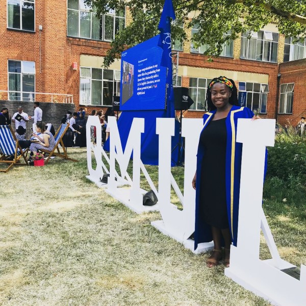 Photo of alumna, Mary Ojo, standing between big white 'QMUL' letters on the lawn outside the Queen's Building at her graduation ceremony