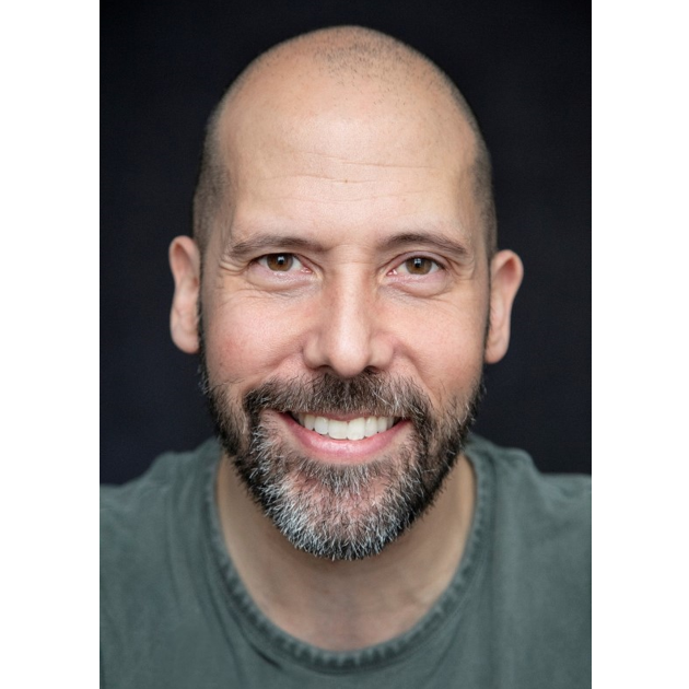 Headshot of alumnus, Simon Nader. He is smiling and wearing a grey t-shirt. The photo is a studio style shot with a black background.