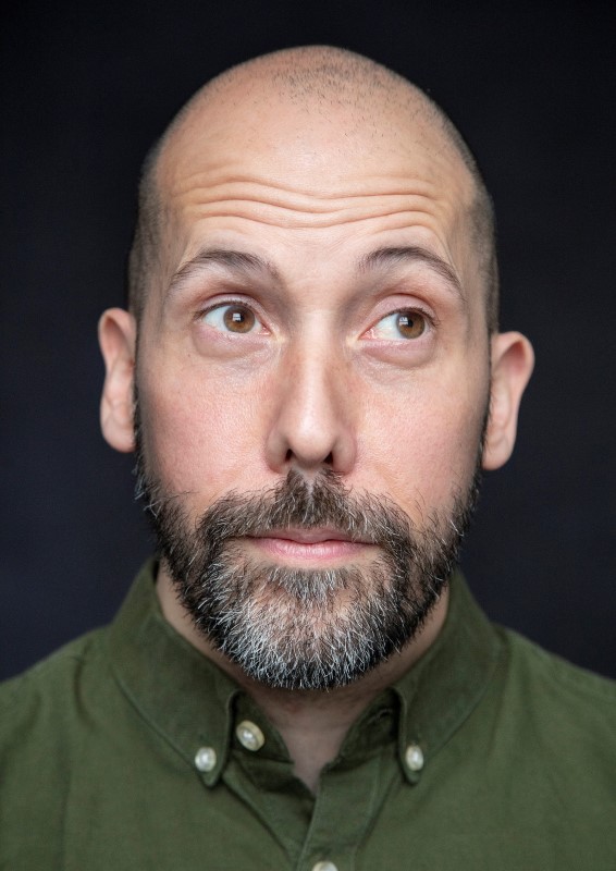 Headshot of alumnus, Simon Nader. He is looking up to the right with a comedic innocent expression. He is wearing a green shirt against a black background.