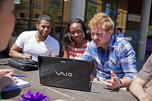 Three students around a laptop outside on a picnic table on campus
