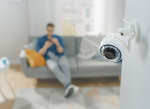 Close up shot of a home security camera on a wall in an apartment. A man is sitting on a sofa in the background. Credit: gorodenkoff/ iStock.com
