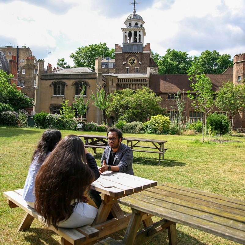 3 students sitting at a picnic table on Charterhouse Square lawn in front of old brick buildings
