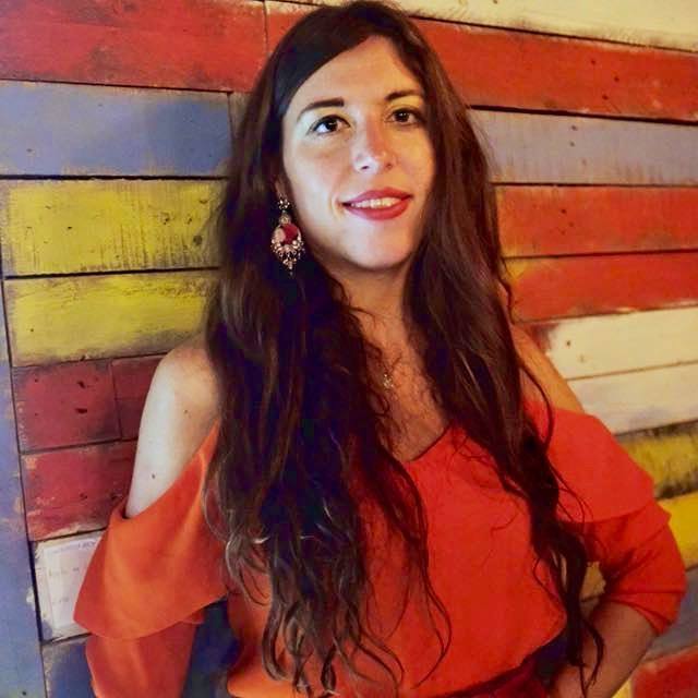 A white woman with long brown hair wears a red dress in front of a colourful wall