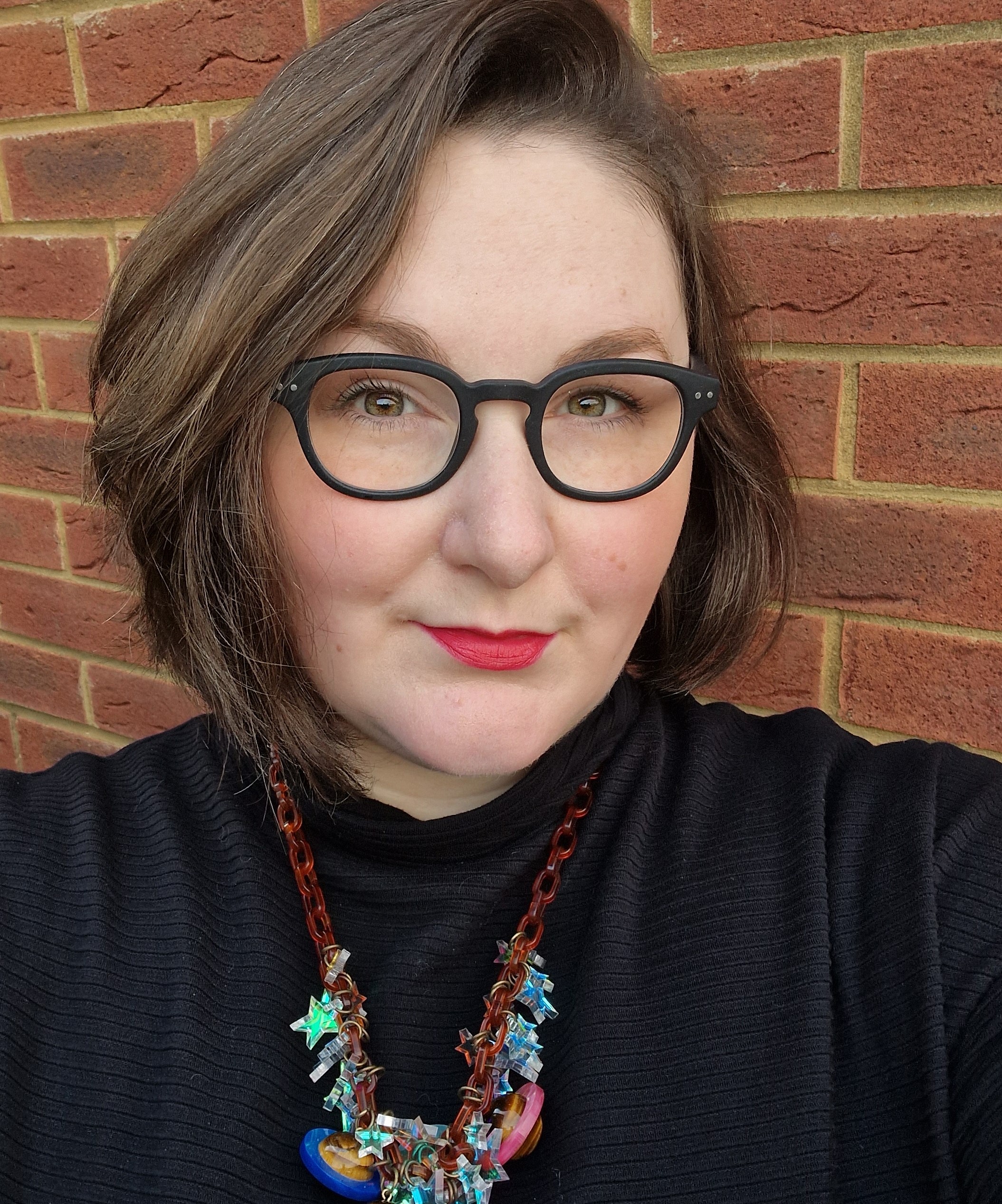 Headshot of Kimberley Freeman, a white woman with glasses wearing a black turtleneck, standing in front of a red brick wall