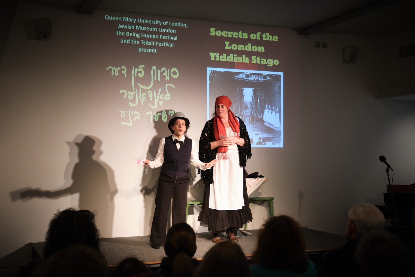 Two actors stand on a stage in front of a projected image saying 'Secrets of the London Yiddish Stage'. One wears a top hat, waistcoat and bow tie, whilst the other wears a shawl, headscarf and apron.