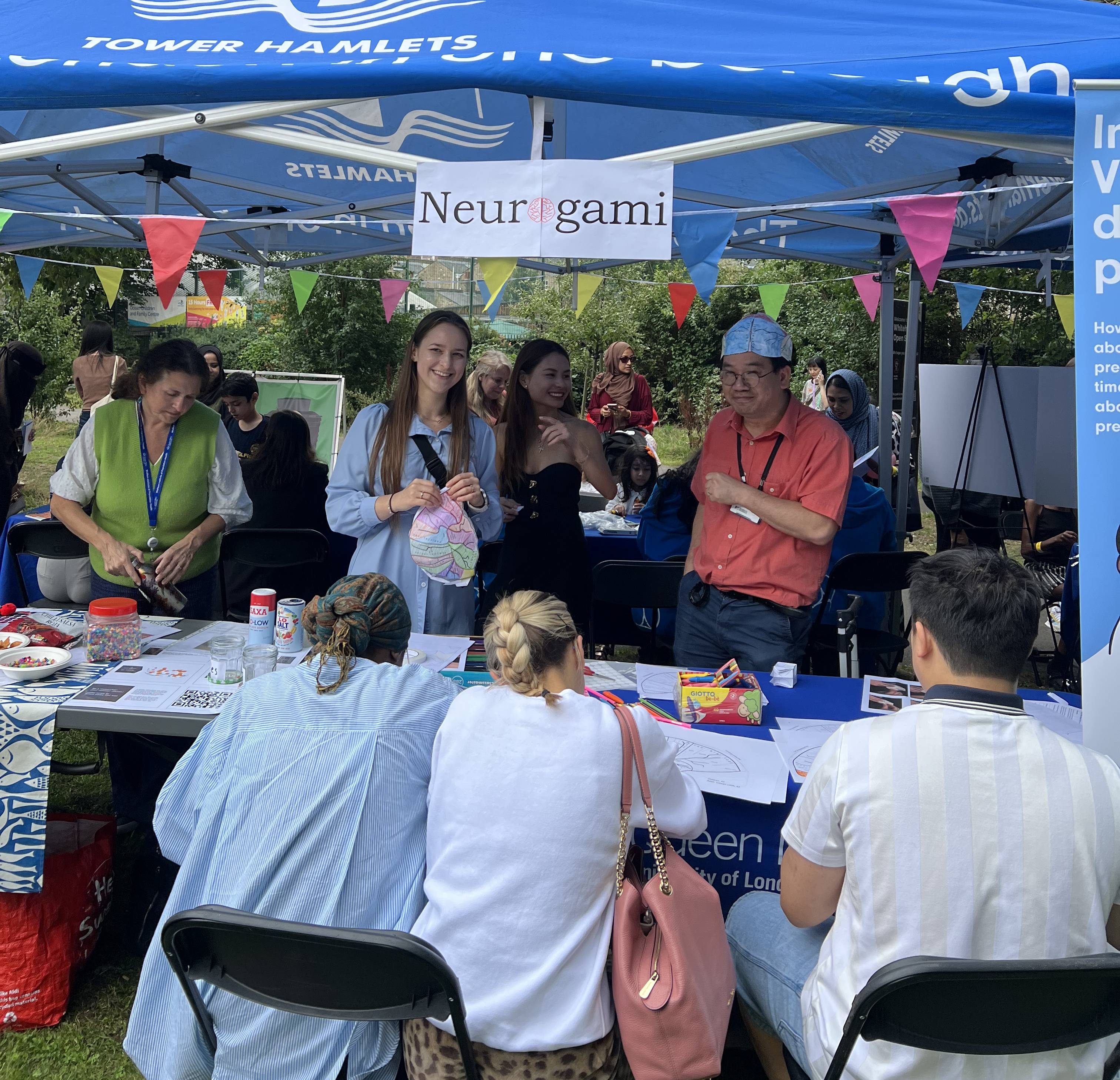 In a blue marquee in a park, smiling people make and colour in paper hats in the shape of brains