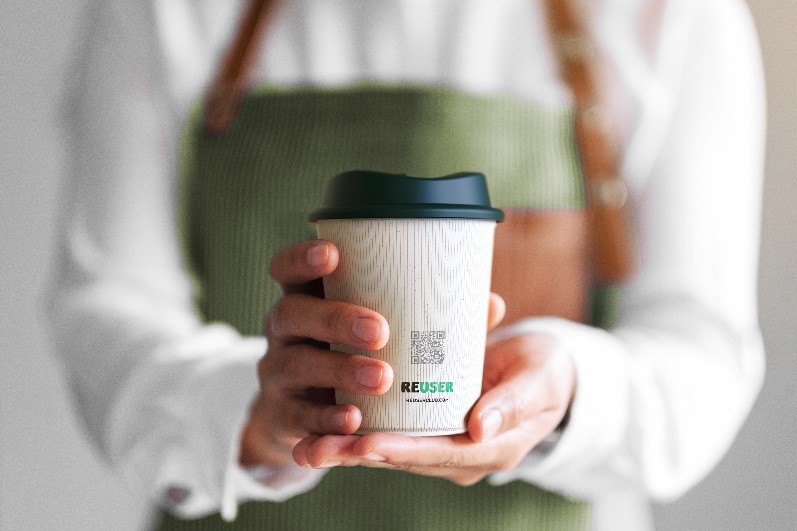 A person in a green apron holding a reusable Reuser coffee cup.