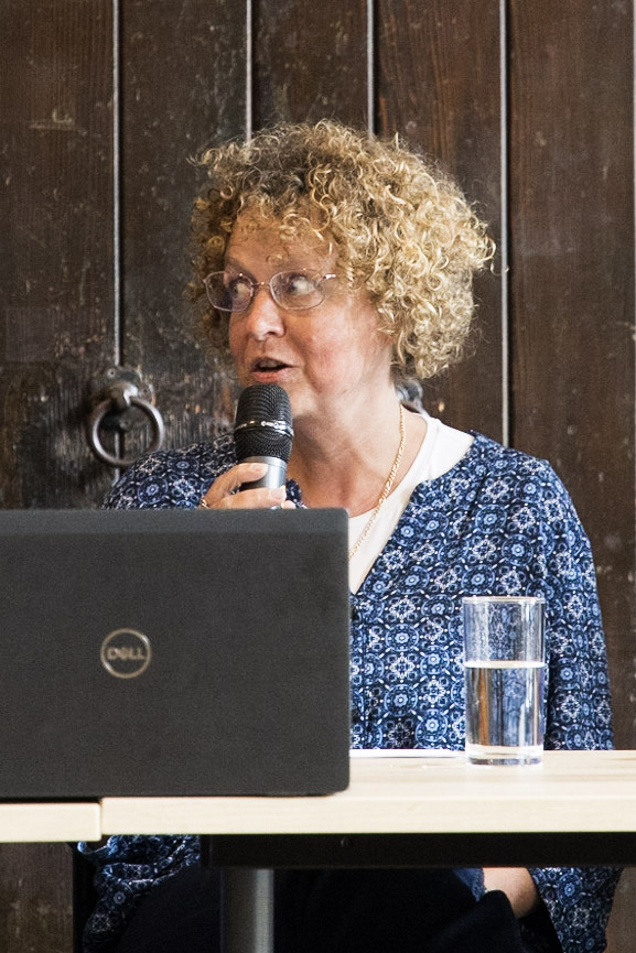 Dr Nicole Brown speaking at the Mile End Institute-DPC event on 22 May. She is speaking into a microphone looking to her right and is sitting behind a panel against a dark wooden background