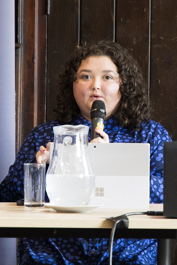 Bethany Bale speaking at the Mile End Institute's event on 22 May. She is holding a microphone, sitting behind a laptop and is looking directly at the camera.
