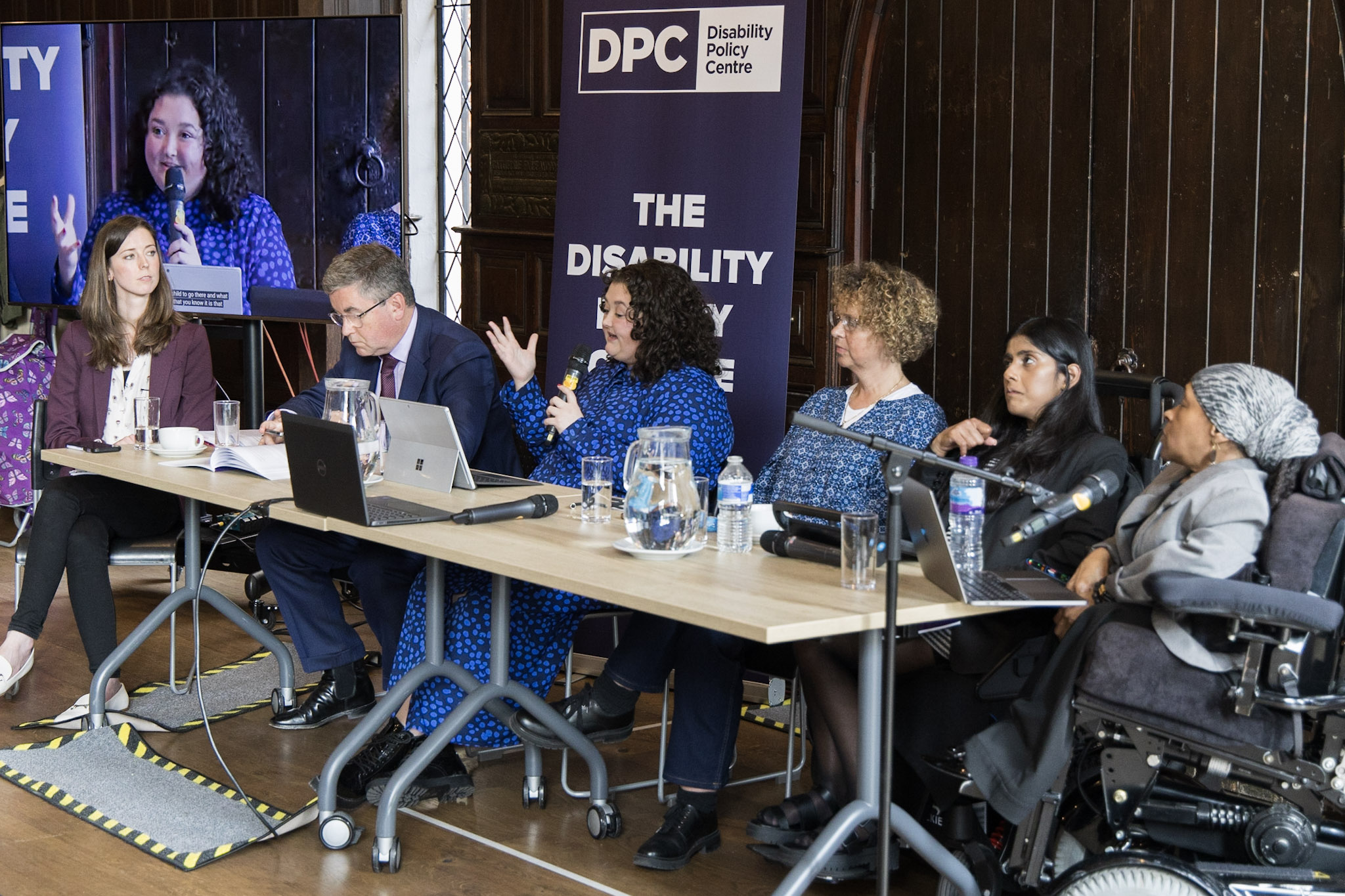 Panel of speakers at the MEI-DPC event on 22 May. From left-to-right, it shows Chloe Schendel-Wilson, Sir Robert Buckland, Bethany Pale, Nicole Brown, Bal Deol, and Michelle Daley sitting at a table against a dark wooden background in the Lecture Hall at Toynbee Hall.