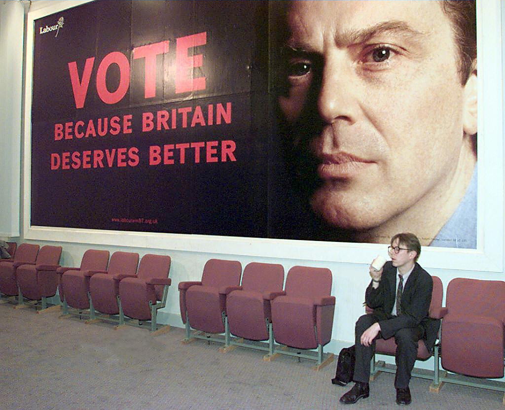 Photo of a man sitting below a poster of Tony Blair urging Britons to vote in 1997.