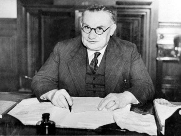 Black and White photo of Ernest Bevin, then Foreign Secretary, working at his desk. He is sitting in front of a pile of papers, wearing a dark suit with a waistcoat, and small round glasses.