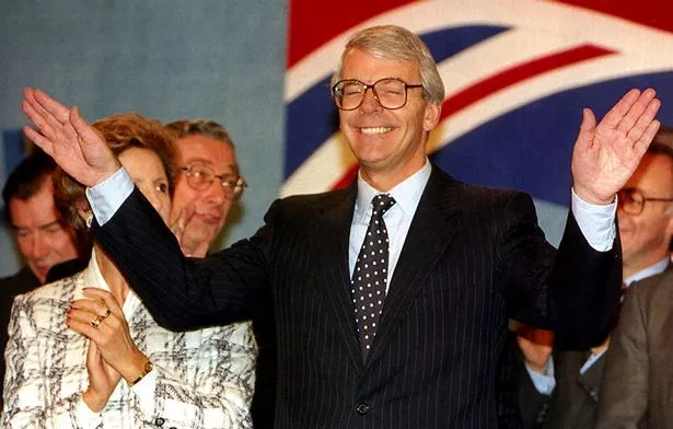 John Major at the Conservative Party conference in Blackpool in October 1993. He is wearing a dark suit, has his arms outstretched and his eyes closed, and is smiling. He is surrounded by the Cabinet and his wife, Norma.