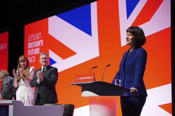 Rachel Reeves giving a speech at the Labour Party's conference in Liverpool in October 2023. She is standing in front of a Union Jack background and Keir Starmer and Angela Rayner can be seen to the left of her, applauding.
