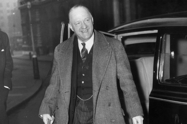 Black and white photo of Rab Butler getting out of a car in Downing Street. He is wearing a three-piece suit and a heavy tweed overcoat and is looking to the cameramen on his left.
