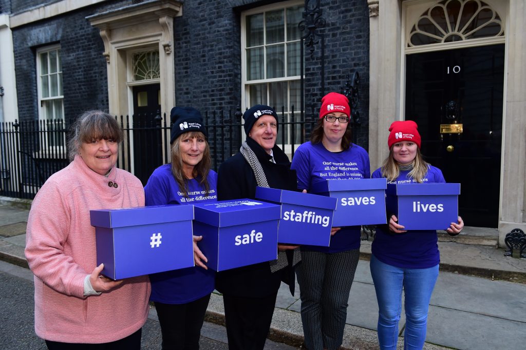 Members of the Royal College of Nursing delivering a petition to Downing Street in February 2020. There are 5 women standing outside the front door to Number 10 and they are holding blue ballot boxes that say '# safe staffing saves lives'.
