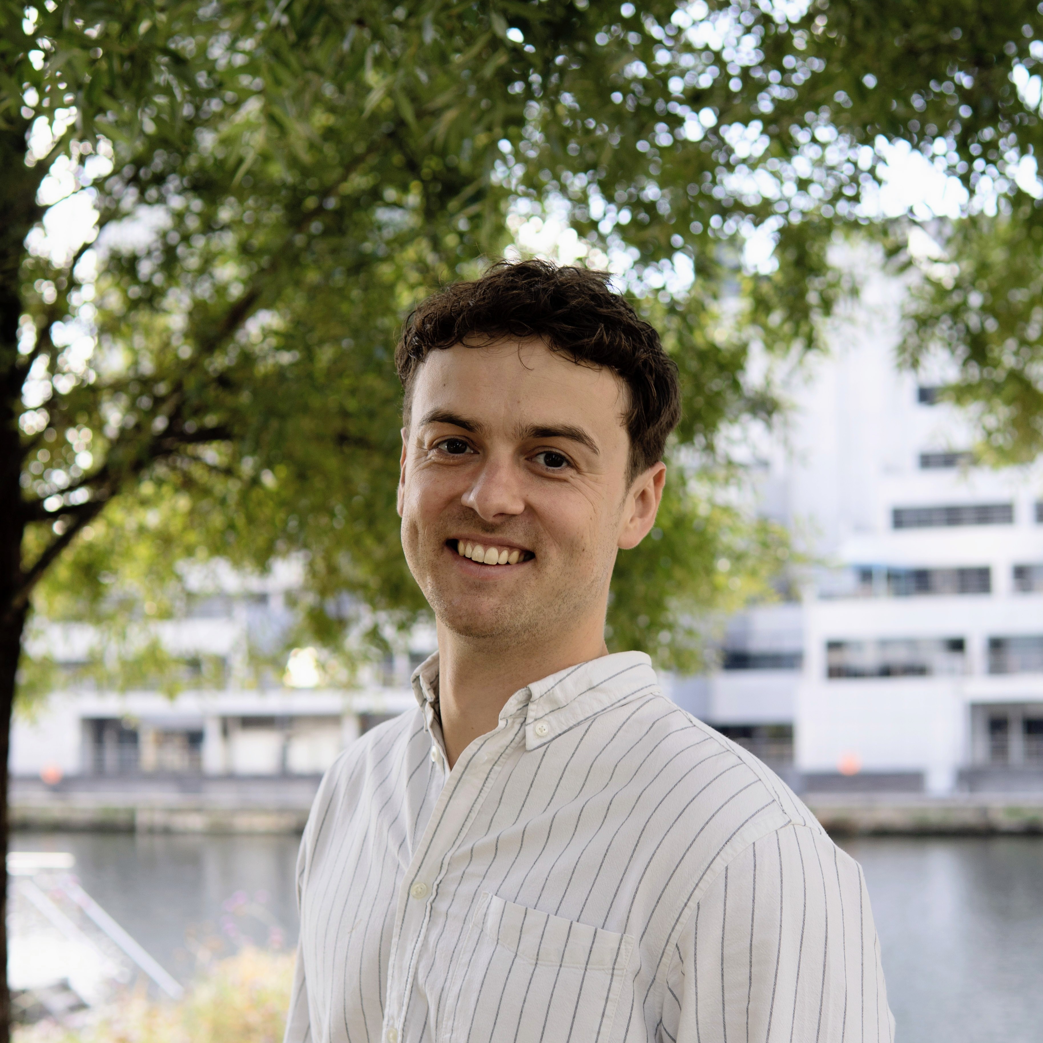 Photo of Dr Matthew Barnfield. He is standing sideways on, wearing a white shirt, and smiling towards the camera.