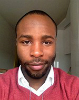 A close-up of Damola looking directly into the camera. He wears a white collared shirt with a red jumper. He has dark skin, short dark hair and a close-shaven, neat beard.