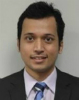 A close-up head and shoulders shot of Syed. He is wearing a suit with a blue striped tie. He has short, neat dark hair and brown eyes. He is clean shaven. He is smiling slightly.