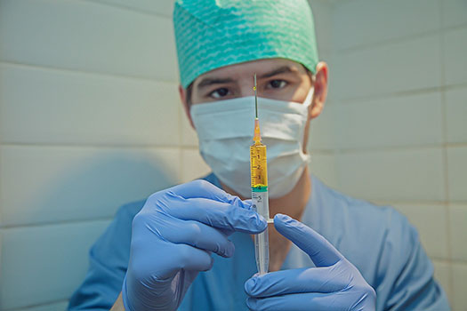 Male nurse wearing a medical mask, scrubs and cap holding a syringe