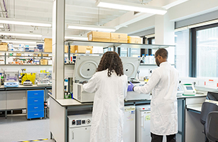 male and female student in lab coats stood by machinery in lab