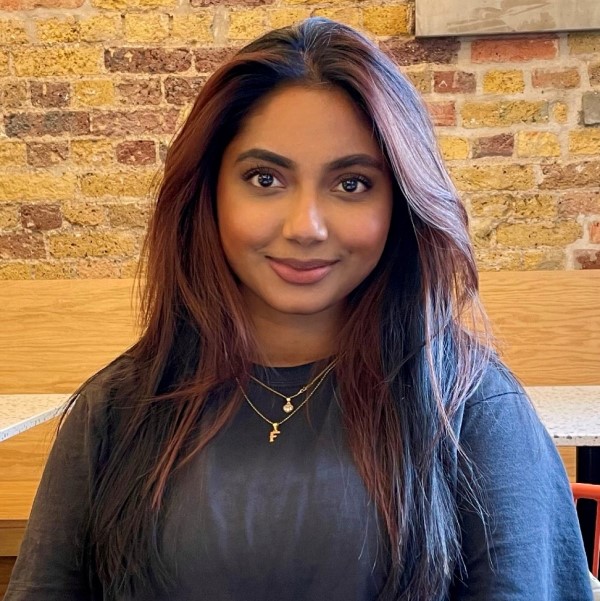 A head and shoulders photo of alumna, Melissa Mahmud. She is wearing a grey long-sleeved top and is sat in front of an indoor brick wall and is smiling at the camera.