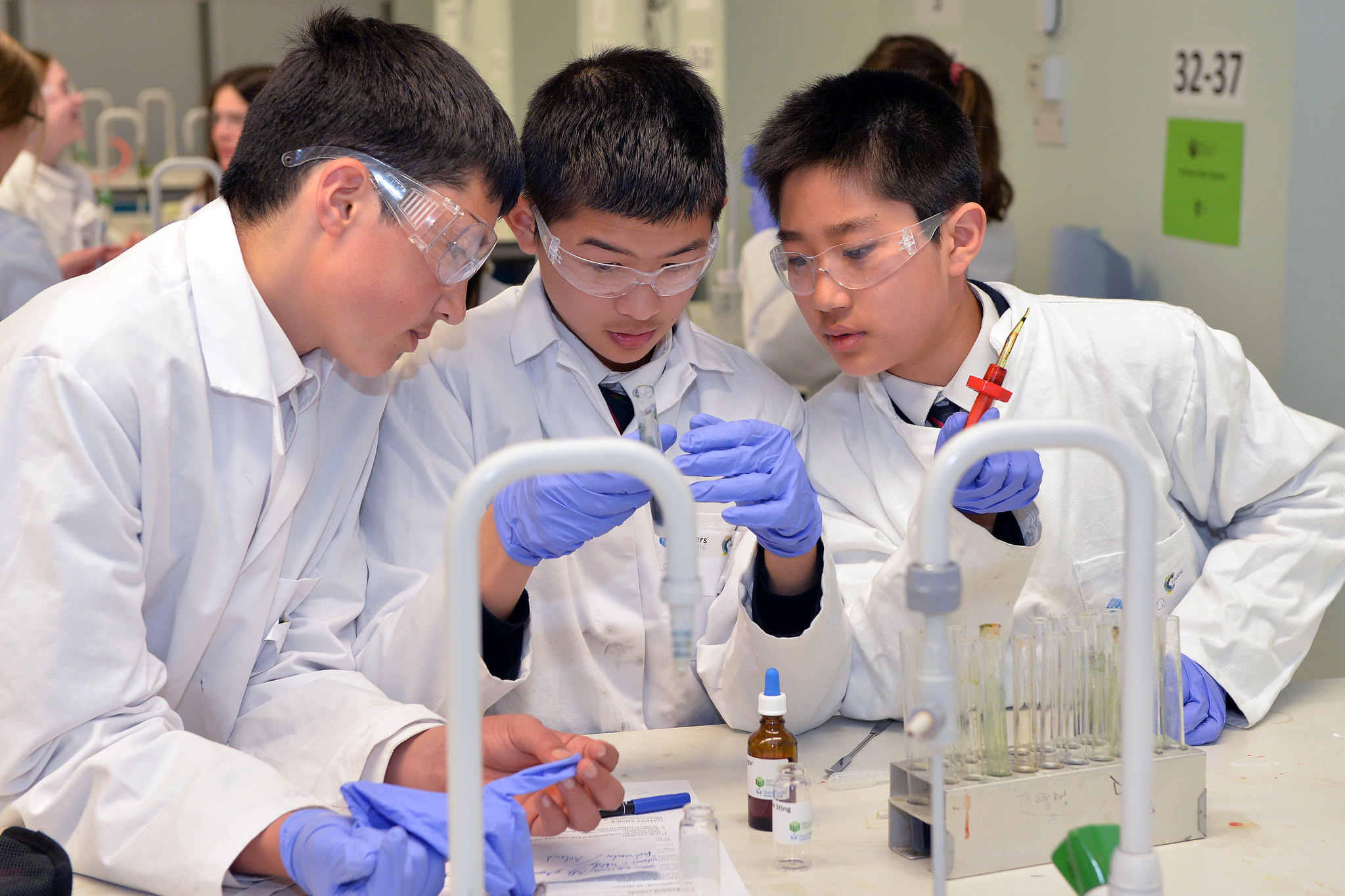 Three school children in lab coats look at a test tube full of a dark liquid