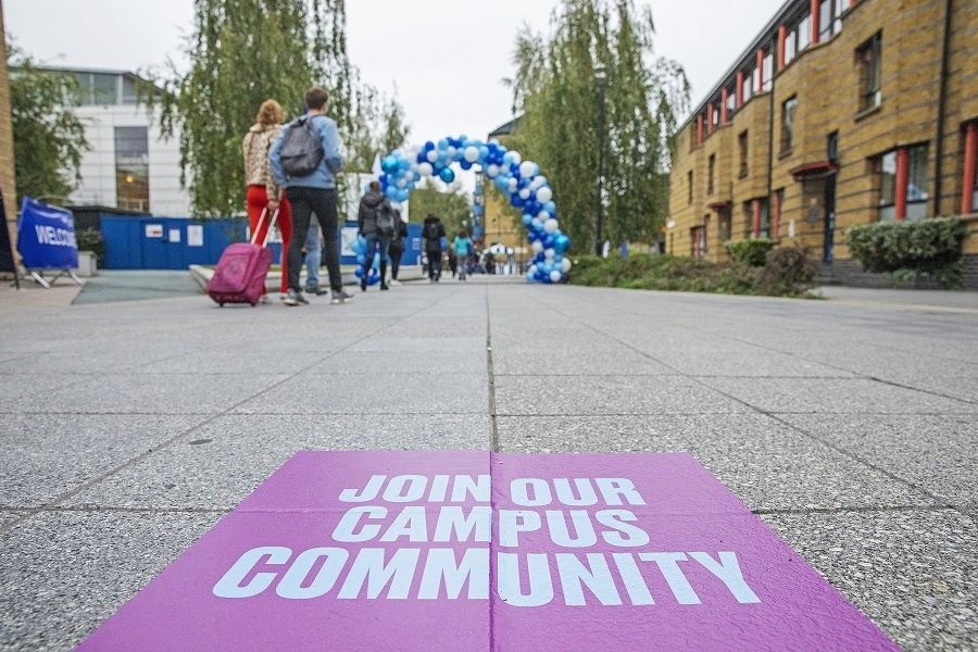 The Mile End Campus showing a student with a suit case and welcome sign on the floor saying 'Join us in our campus community'