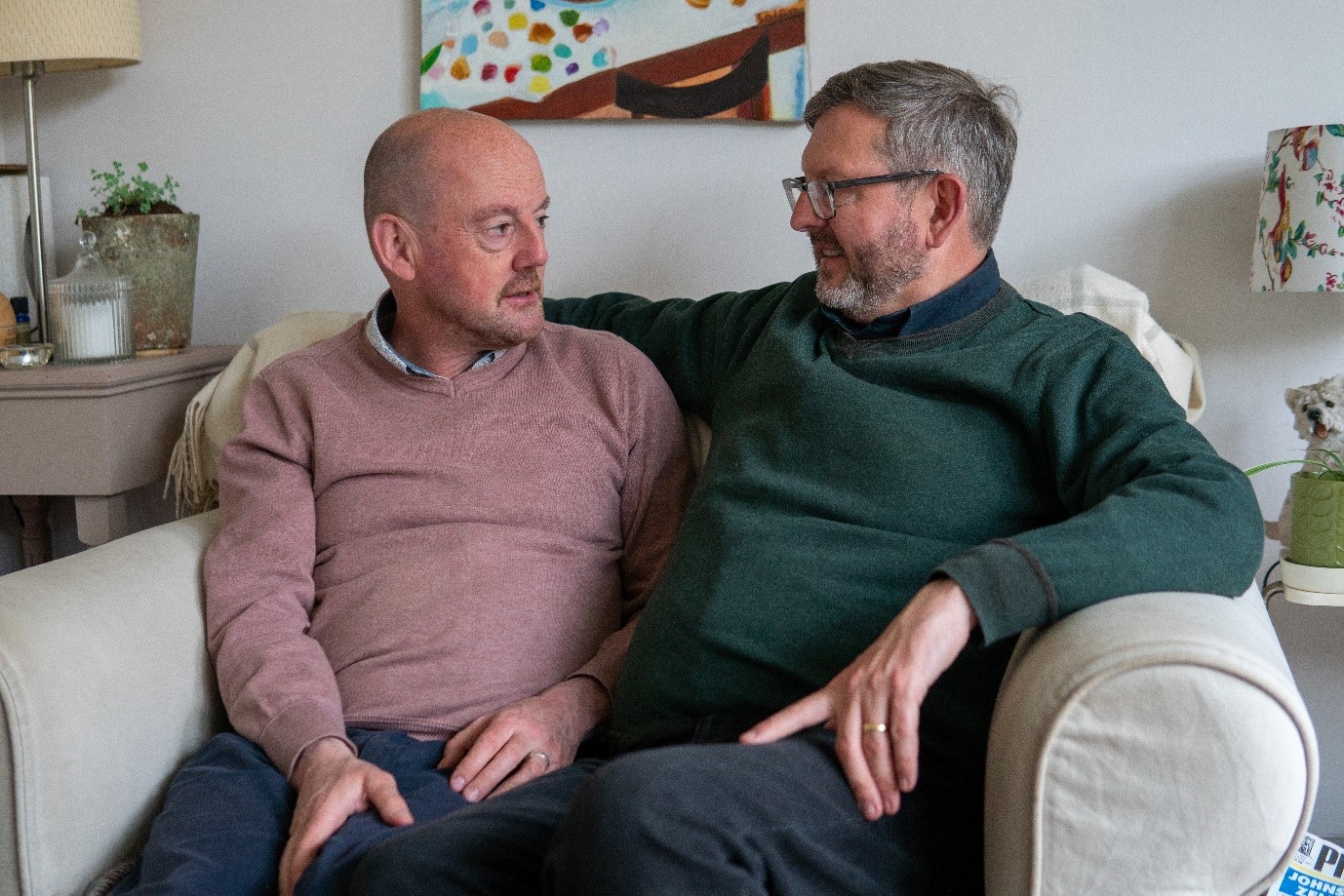Two older men, a married couple, sit closely together on their sofa, in conversation. One is reassuring the other by putting his arm around him.
