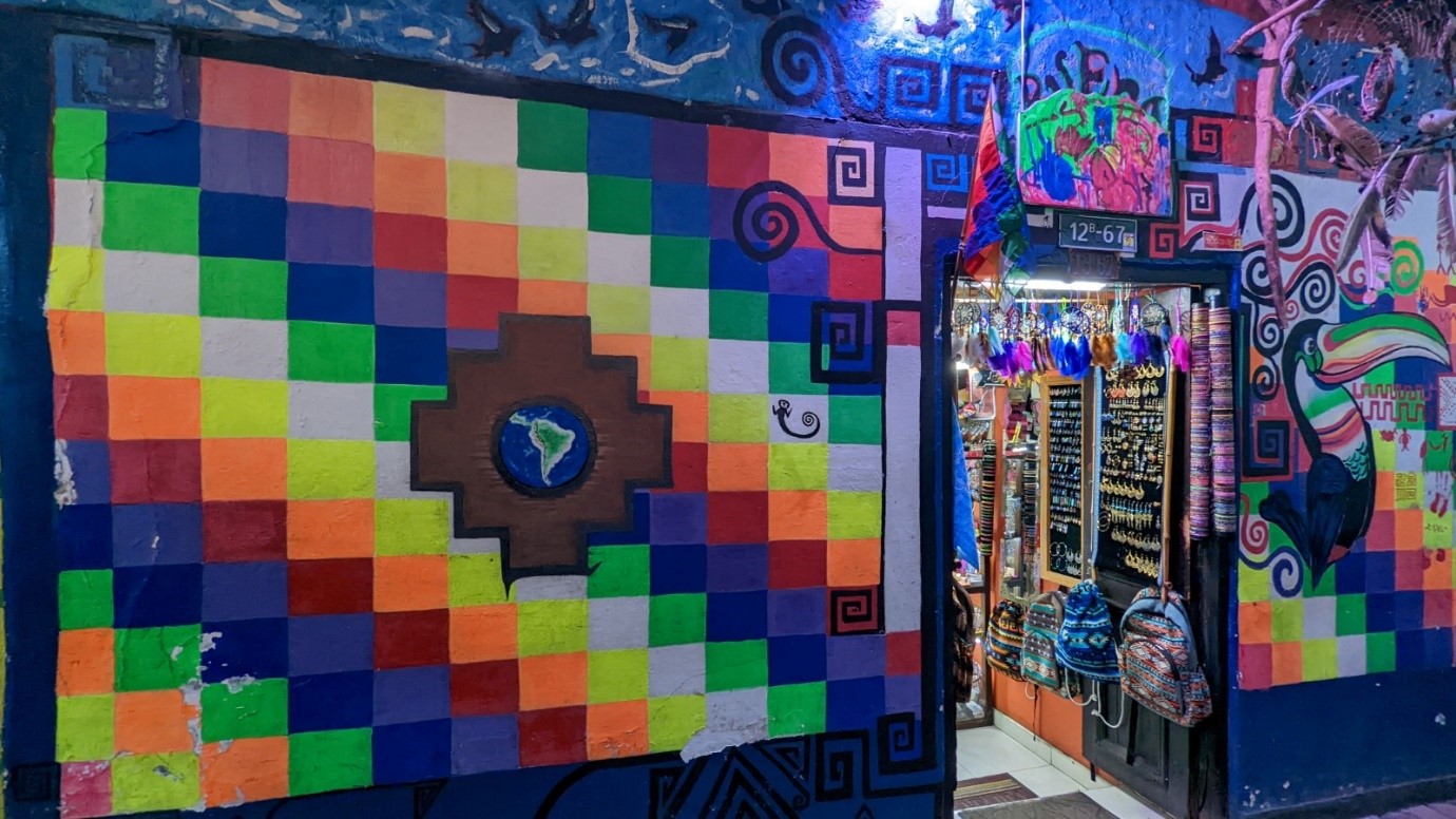 The photo shows a mural painted on the walls either side of a shop doorway. On one side is painted the flag of the indigenous people of Latin America, which consists of diagonal stripes in many colours with a view of the Earth in the centre of the flag, showing the continent of Latin and South America. The mural continues on the other side of the door with a painting of a toucan. Bags and jewellery are visible through the shop door.